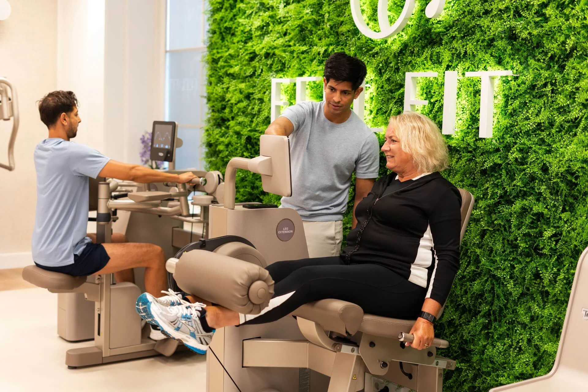 Coach motiveert sporter tijdens fitnessoefening op Technogym-apparaat.